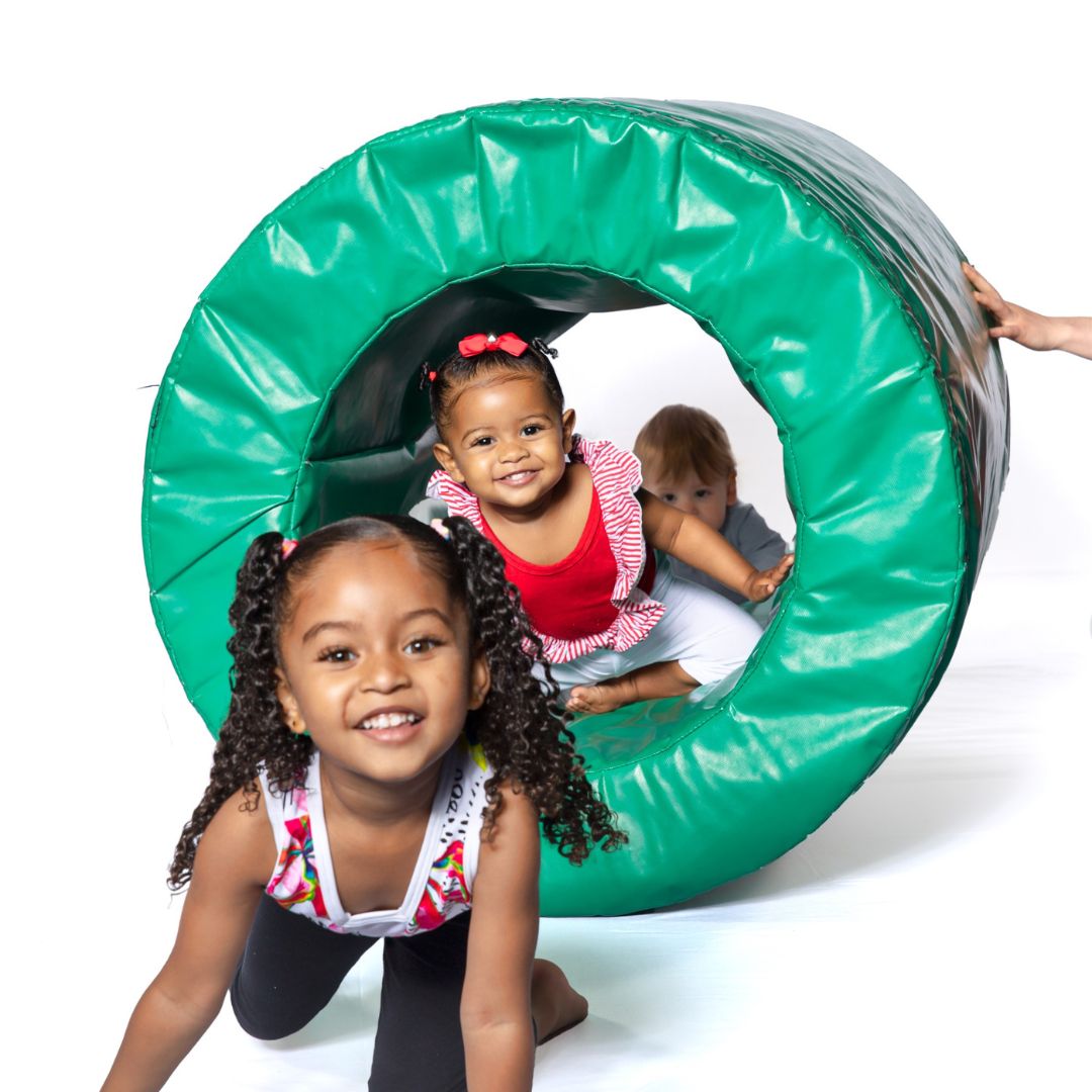 Three young kids crawling through a foam cylinder looking at camera.